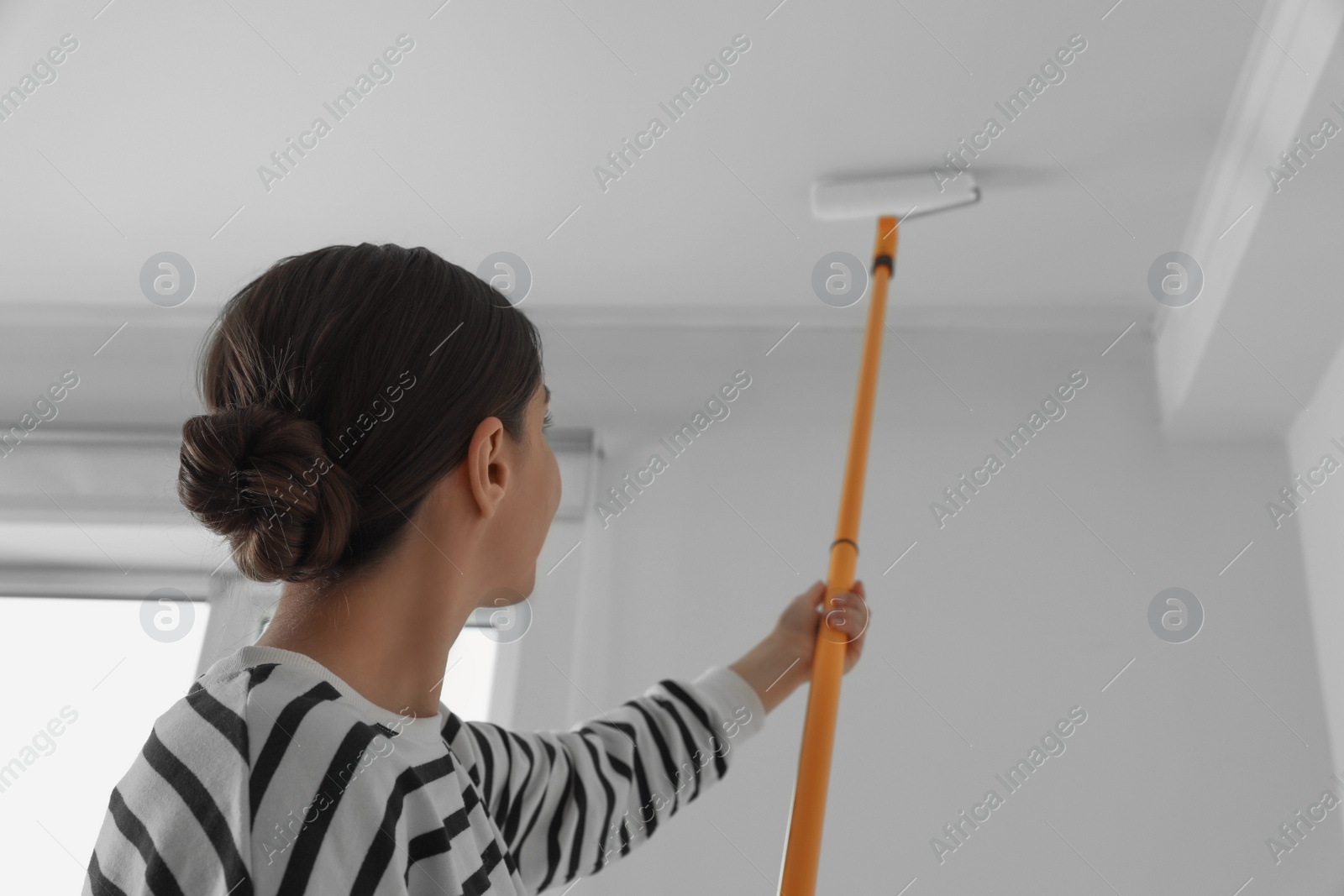 Photo of Young woman painting ceiling with white dye indoors, back view