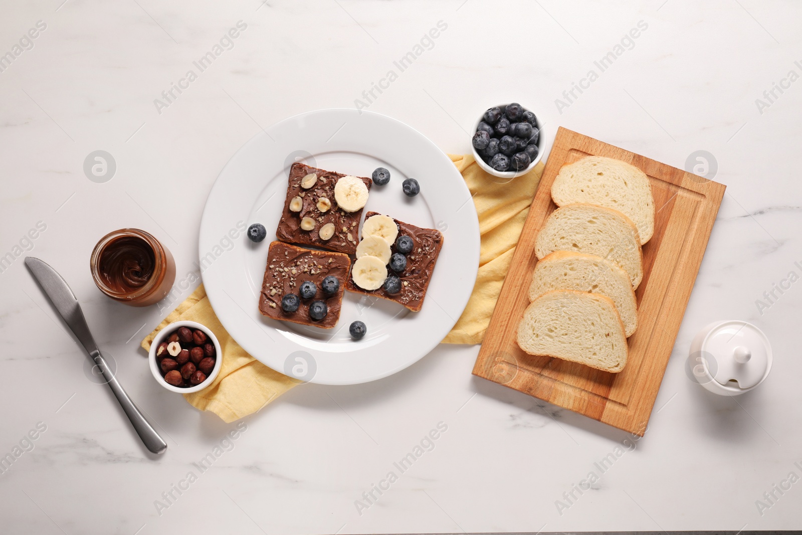 Photo of Toasts with tasty nut butter, banana slices, blueberries and hazelnuts on white marble table, flat lay