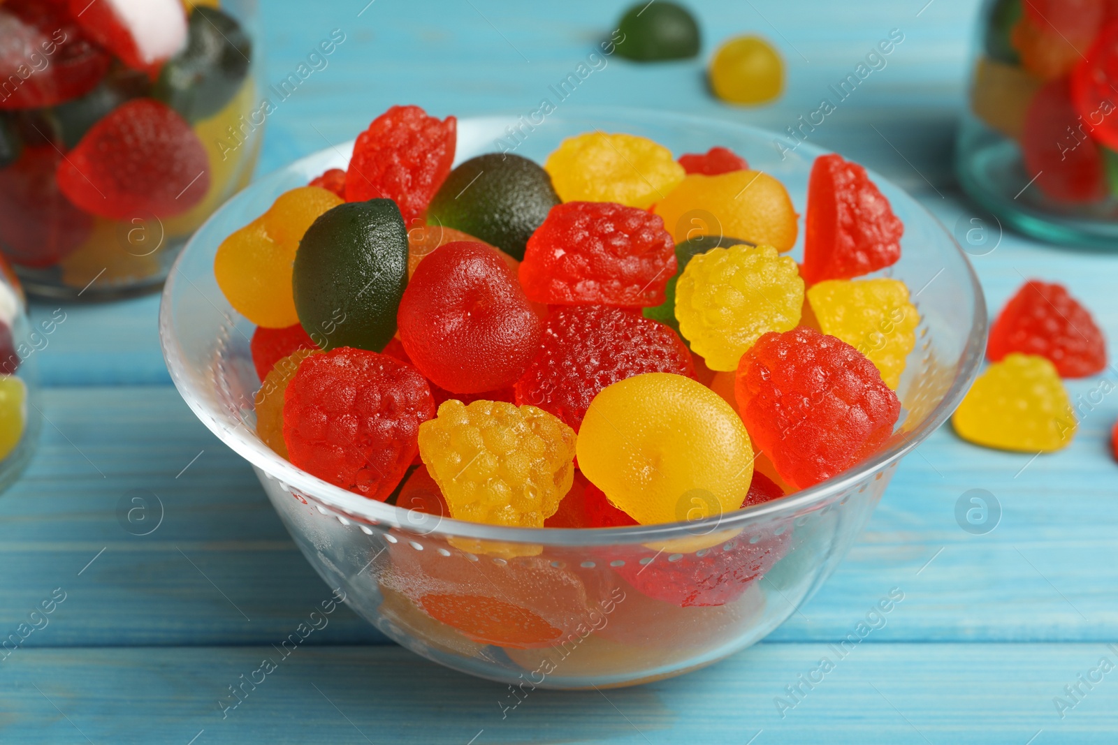 Photo of Delicious gummy fruit shaped candies in glass bowl on light blue wooden table, closeup