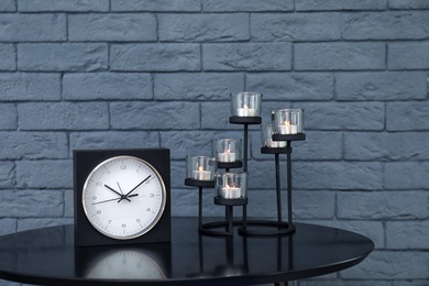 Stylish analog clock and candles on table near brick wall