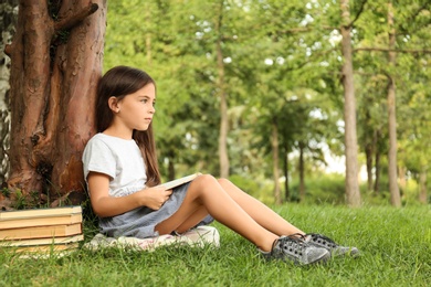 Photo of Cute little girl reading book on green grass near tree in park