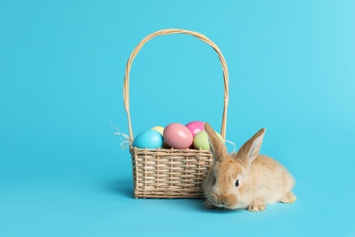 Adorable furry Easter bunny near wicker basket with dyed eggs on color background