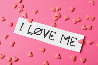 Photo of Piece of paper with handwritten phrase I Love Me and heart shaped sprinkles on pink background, above view