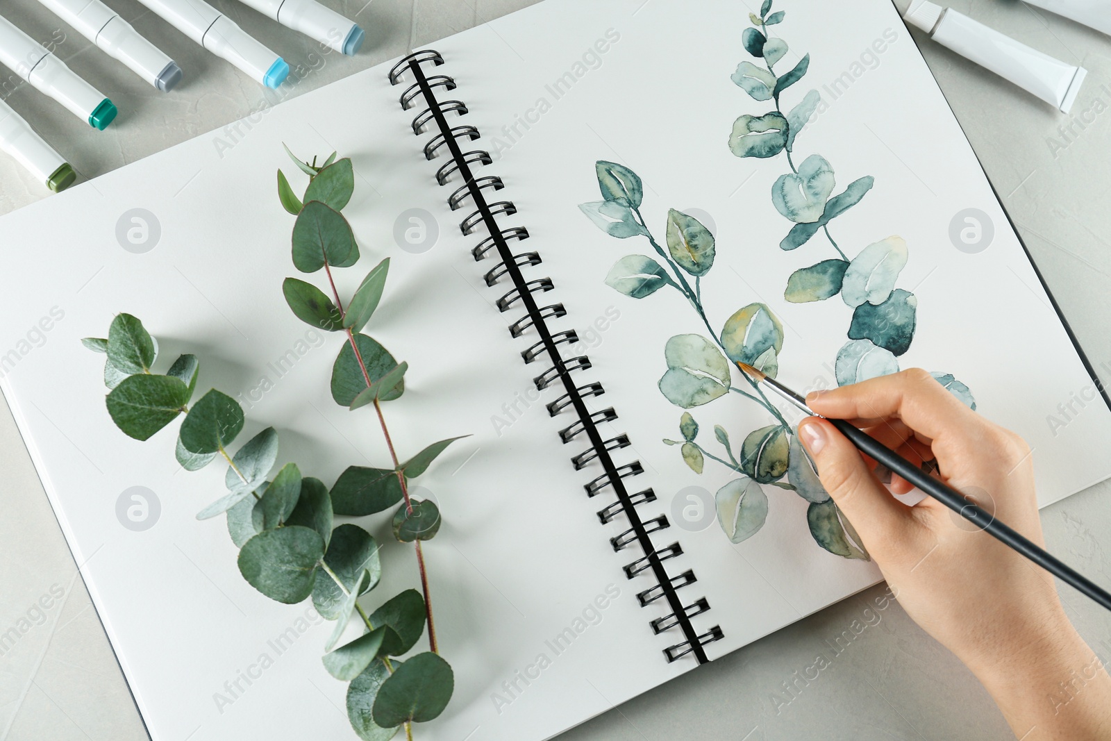 Photo of Woman drawing beautiful eucalyptus branches in sketchbook at white table, top view