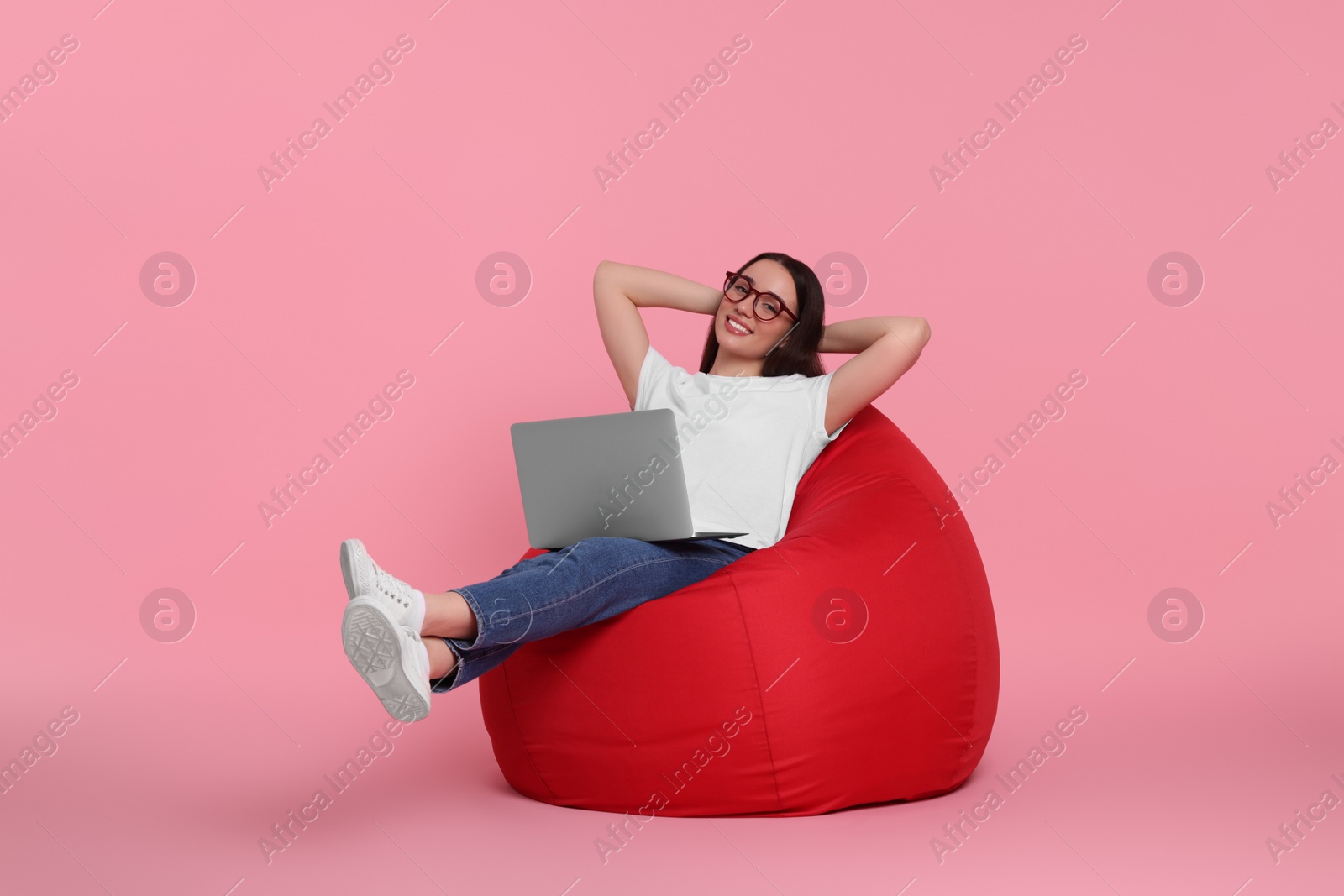 Photo of Smiling young woman with laptop sitting on beanbag chair against pink background