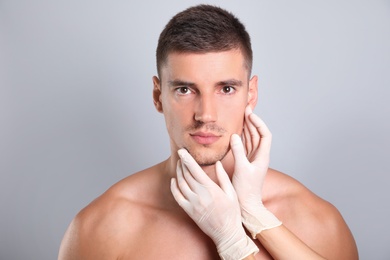 Doctor examining man's face for cosmetic surgery on grey background