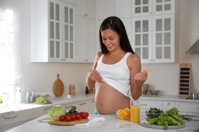 Photo of Young pregnant woman with slice of bell pepper near table in kitchen. Taking care of baby health