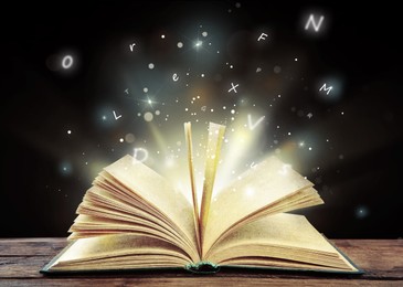 Open book with magic light and glowing letters flying out of it on wooden table against black background