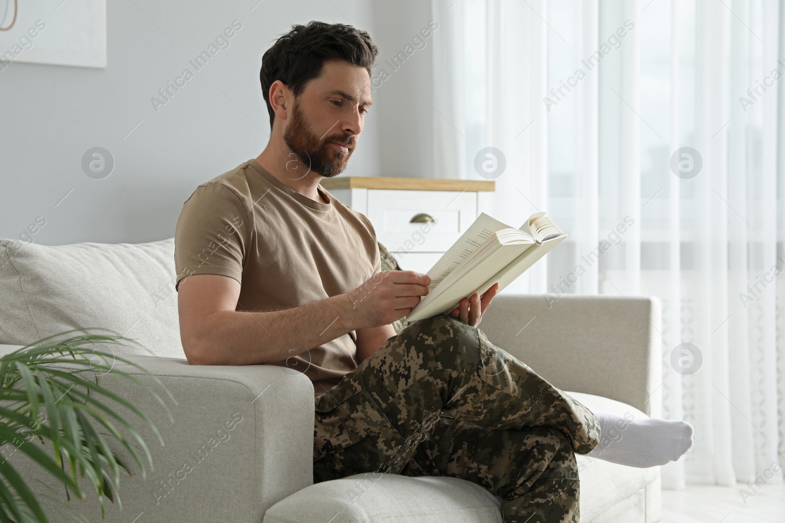 Photo of Soldier reading book on soft sofa in living room. Military service