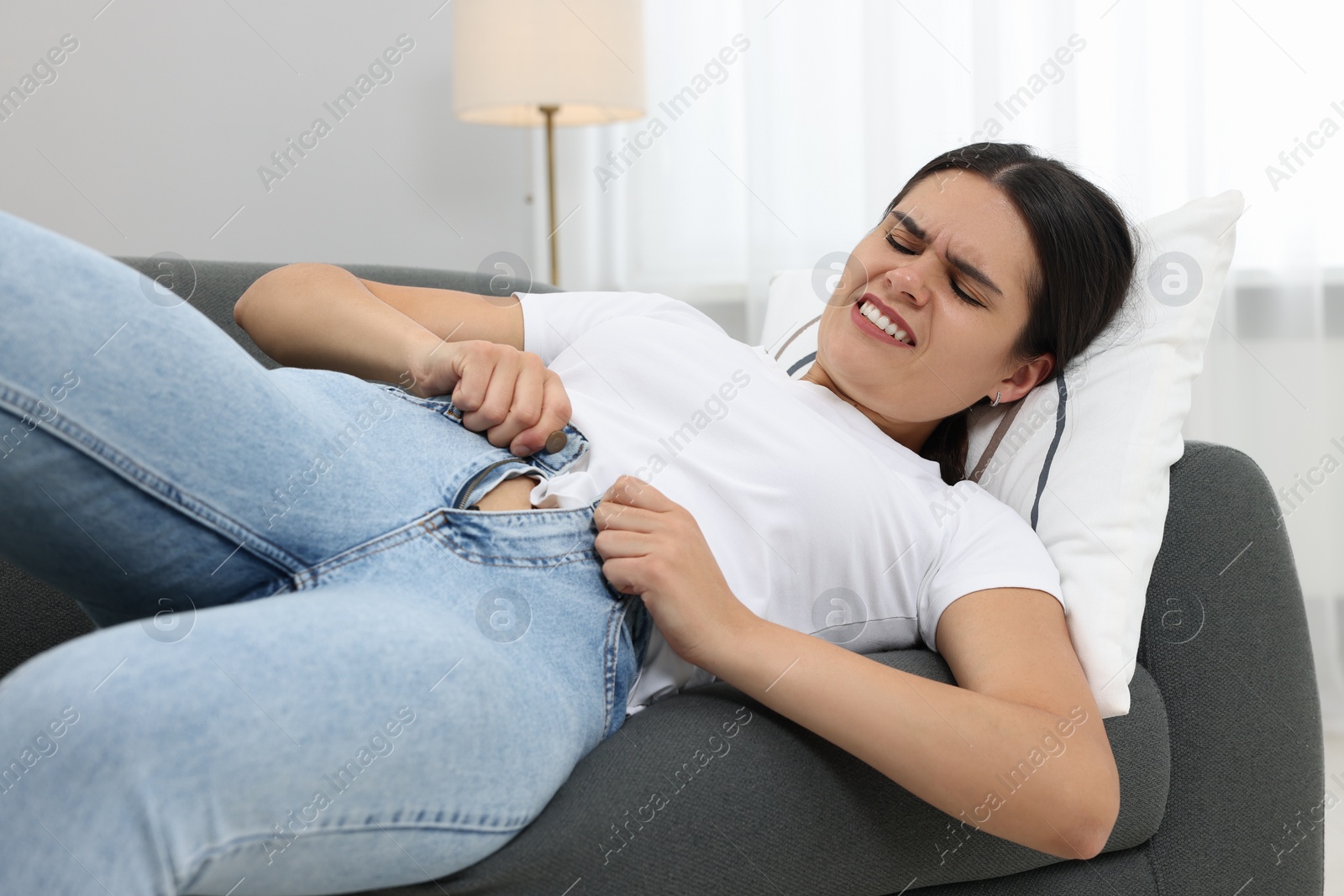 Photo of Young woman struggling to squeeze into tight jeans while lying on sofa at home