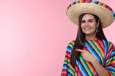 Photo of Young woman in Mexican sombrero hat and poncho pointing at something on pink background. Space for text