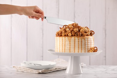 Woman cutting caramel drip cake decorated with popcorn and pretzels at white marble table, closeup