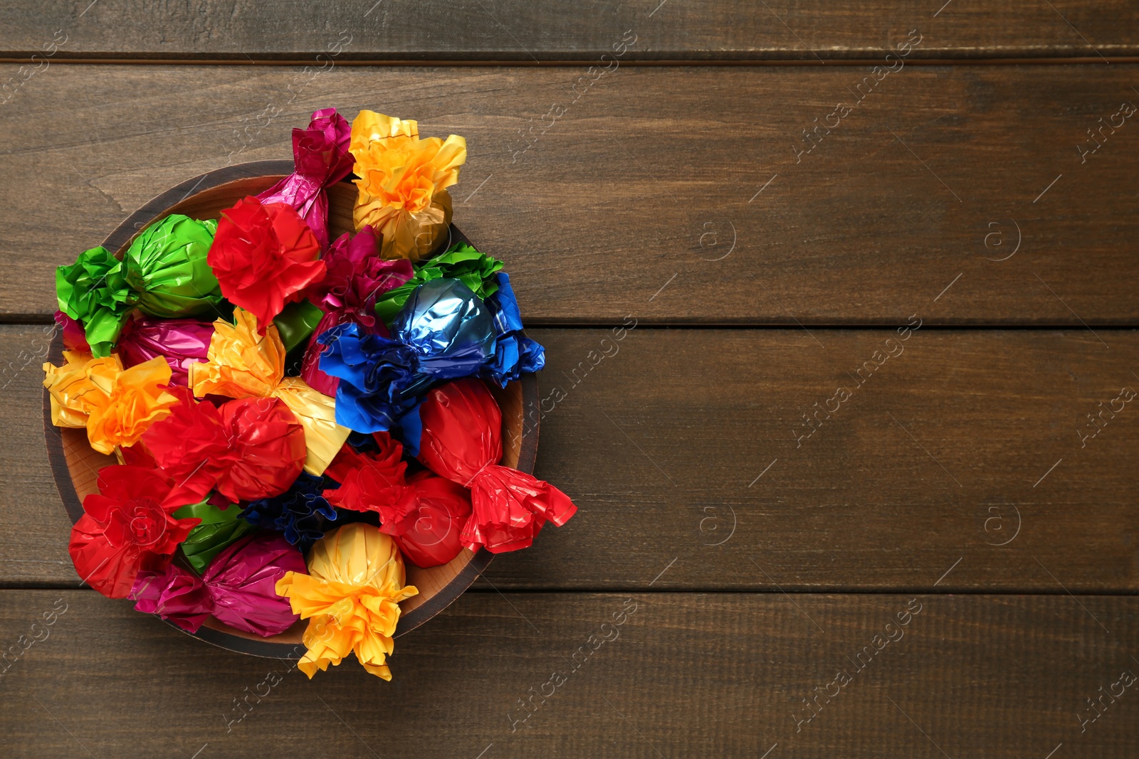 Photo of Bowl with candies in colorful wrappers on wooden table, top view. Space for text