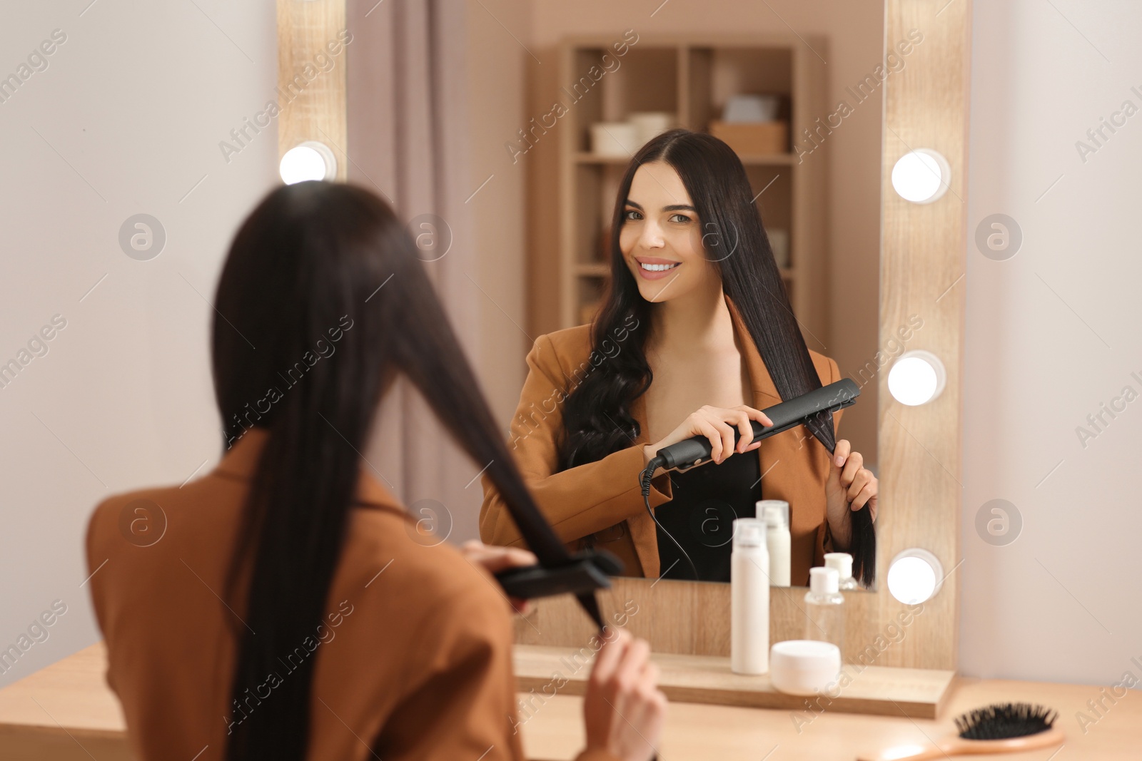 Photo of Beautiful happy woman using hair iron near mirror in room