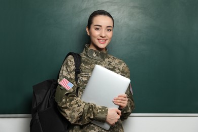 Photo of Female soldier with laptop and backpack near green chalkboard. Military education