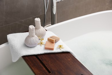 Wooden bath tray with herbal massage bags and bathroom amenities on tub, closeup