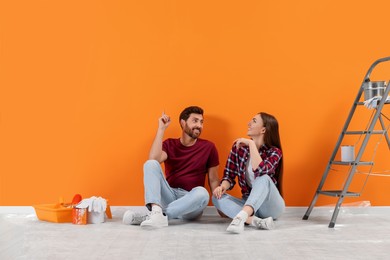 Photo of Man pointing upwards and woman sitting on floor near freshly painted orange wall indoors. Interior design
