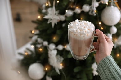 Woman with cup of delicious hot drink near Christmas tree at home, closeup. Space for text