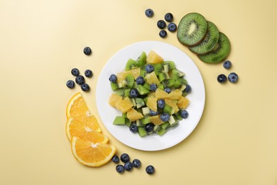 Plate of tasty fruit salad and ingredients on pale yellow background, flat lay