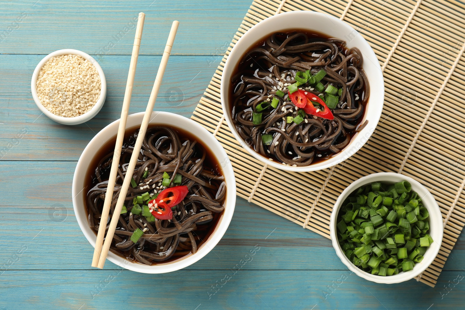 Photo of Tasty buckwheat noodle (soba) soup with chili pepper, green onion, sesame and chopsticks on light blue wooden table, flat lay