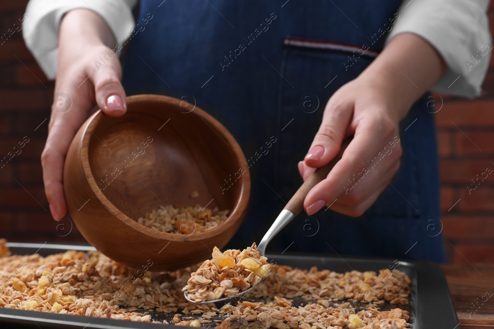 Photo of Woman putting granola from baking tray into bowl at table, closeup