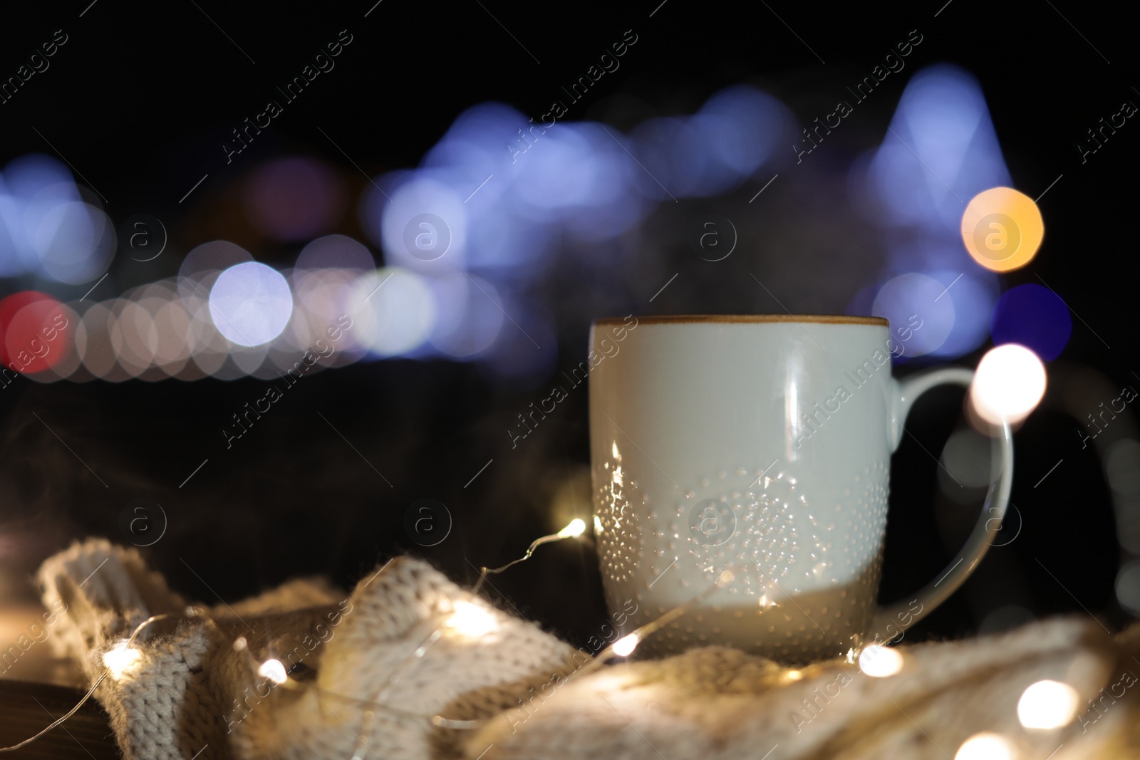 Photo of Cup of hot beverage, sweater and Christmas lights outdoors on winter night. Space for text