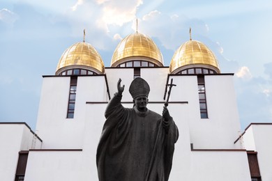 Photo of LVIV, UKRAINE - MAY, 04, 2022: Pope John Paul II statue near Nativity of Holy Virgin Church against cloudy sky, low angle view