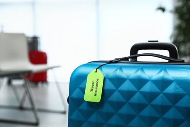 Photo of Blue suitcase with TRAVEL INSURANCE label indoors, closeup. Space for text