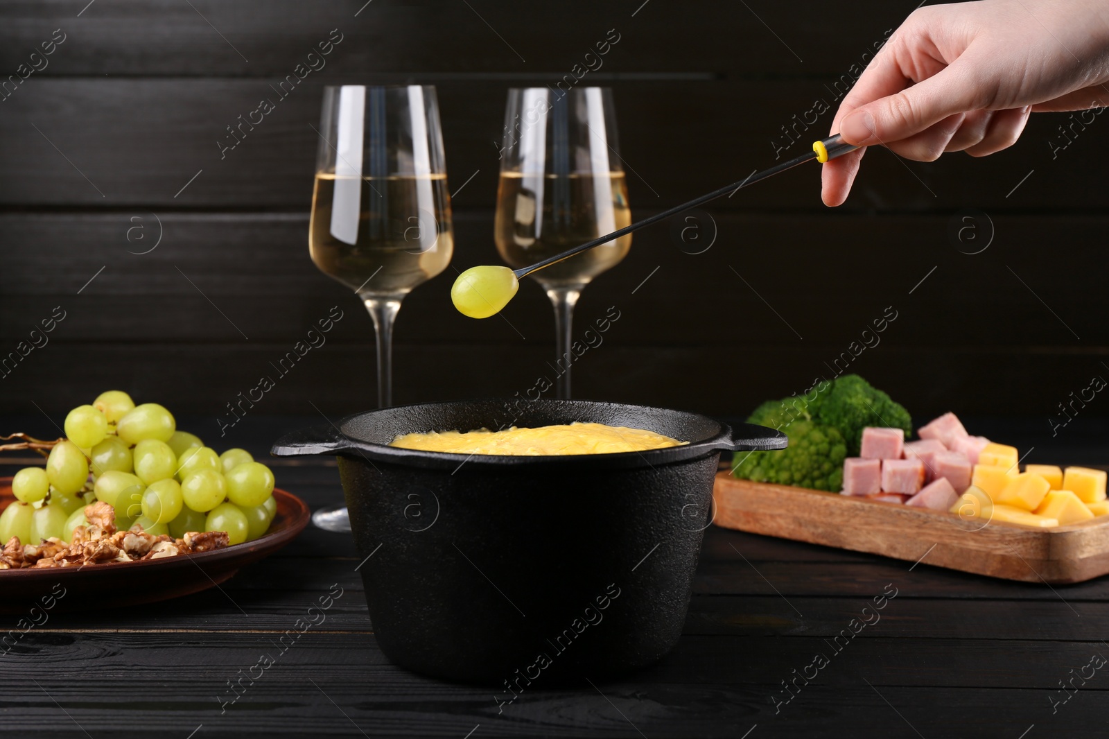 Photo of Woman dipping grape into fondue pot with melted cheese at black wooden table, closeup