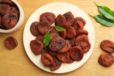 Tasty apricots and green leaves with plate on wooden table, flat lay. Dried fruits