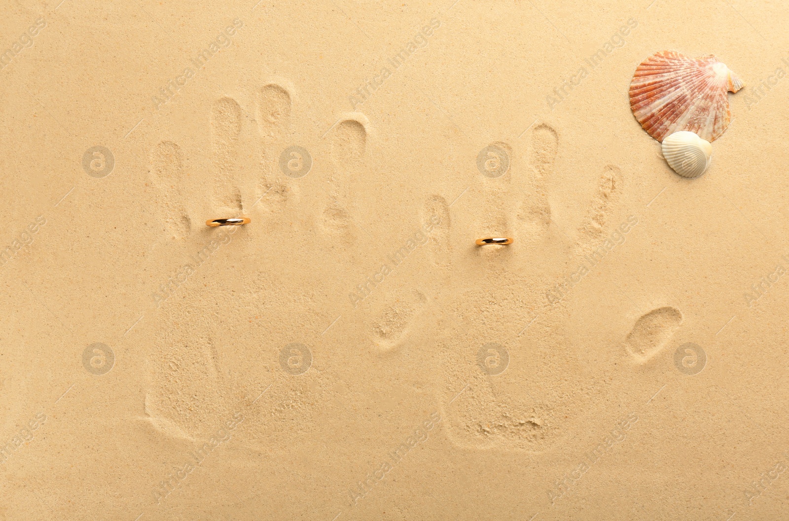 Photo of Honeymoon concept. Handprints, seashells and two golden rings on sand, top view