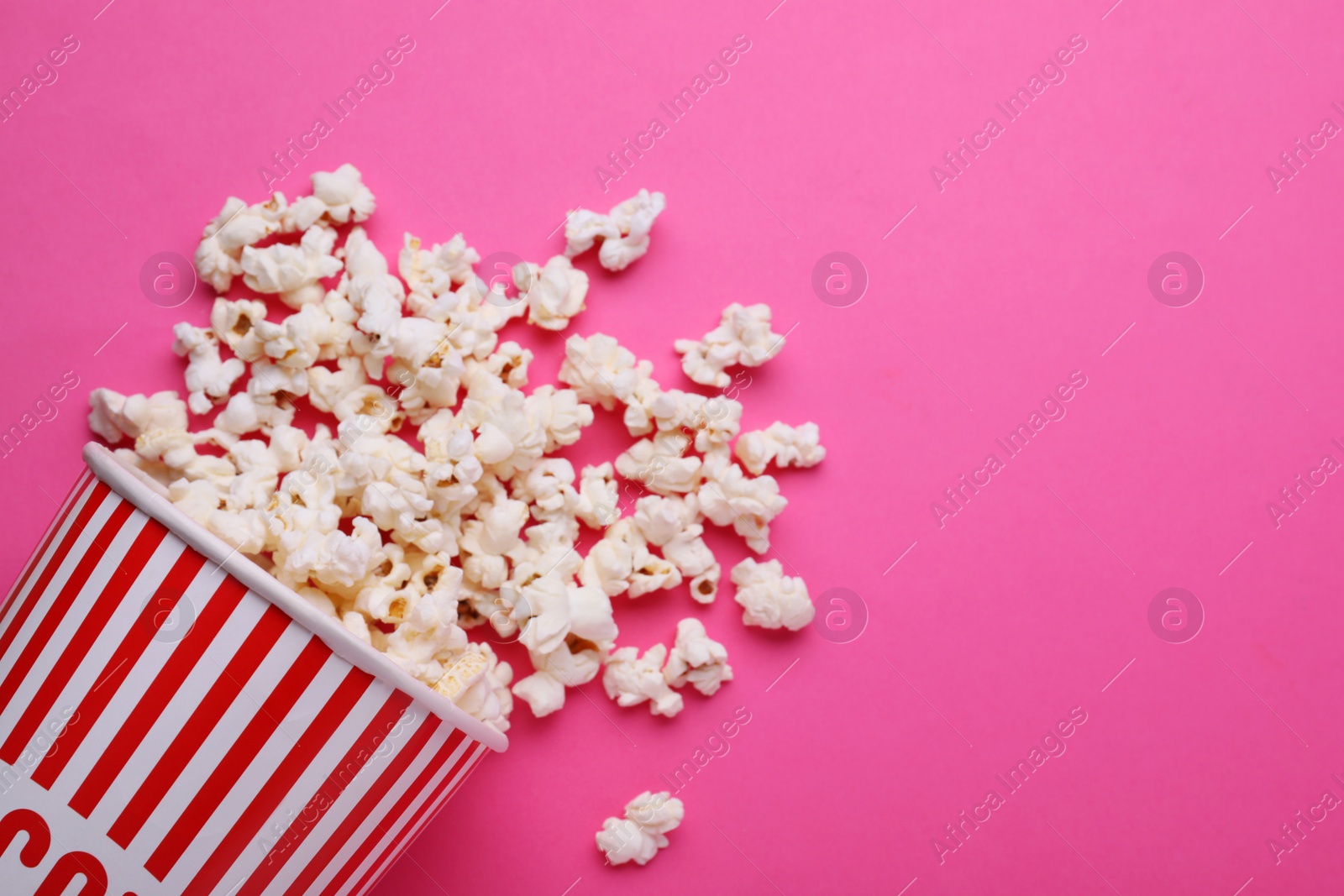 Photo of Overturned paper cup with delicious popcorn on pink background, flat lay. Space for text