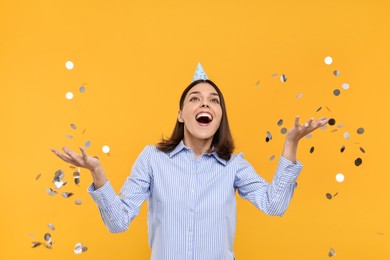 Happy young woman in party hat near flying confetti on yellow background