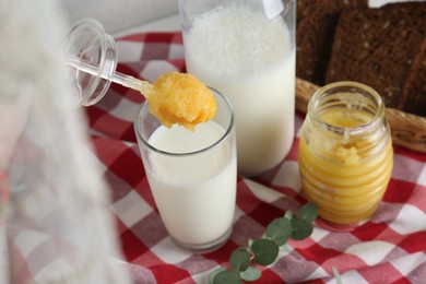 Photo of Putting spoon with tasty honey into glass of milk on checkered cloth, closeup