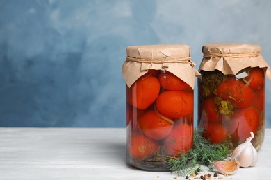 Pickled tomatoes in glass jars and products on white wooden table against blue background, space for text