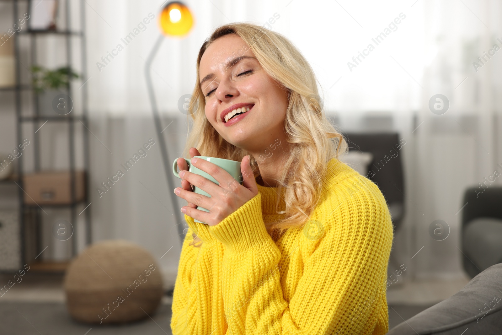 Photo of Happy woman in stylish warm sweater holding cup of drink at home