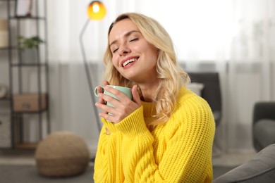 Photo of Happy woman in stylish warm sweater holding cup of drink at home