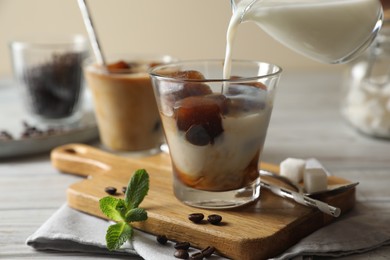 Pouring milk from jug into glass of delicious iced coffee, beans and mint on white table