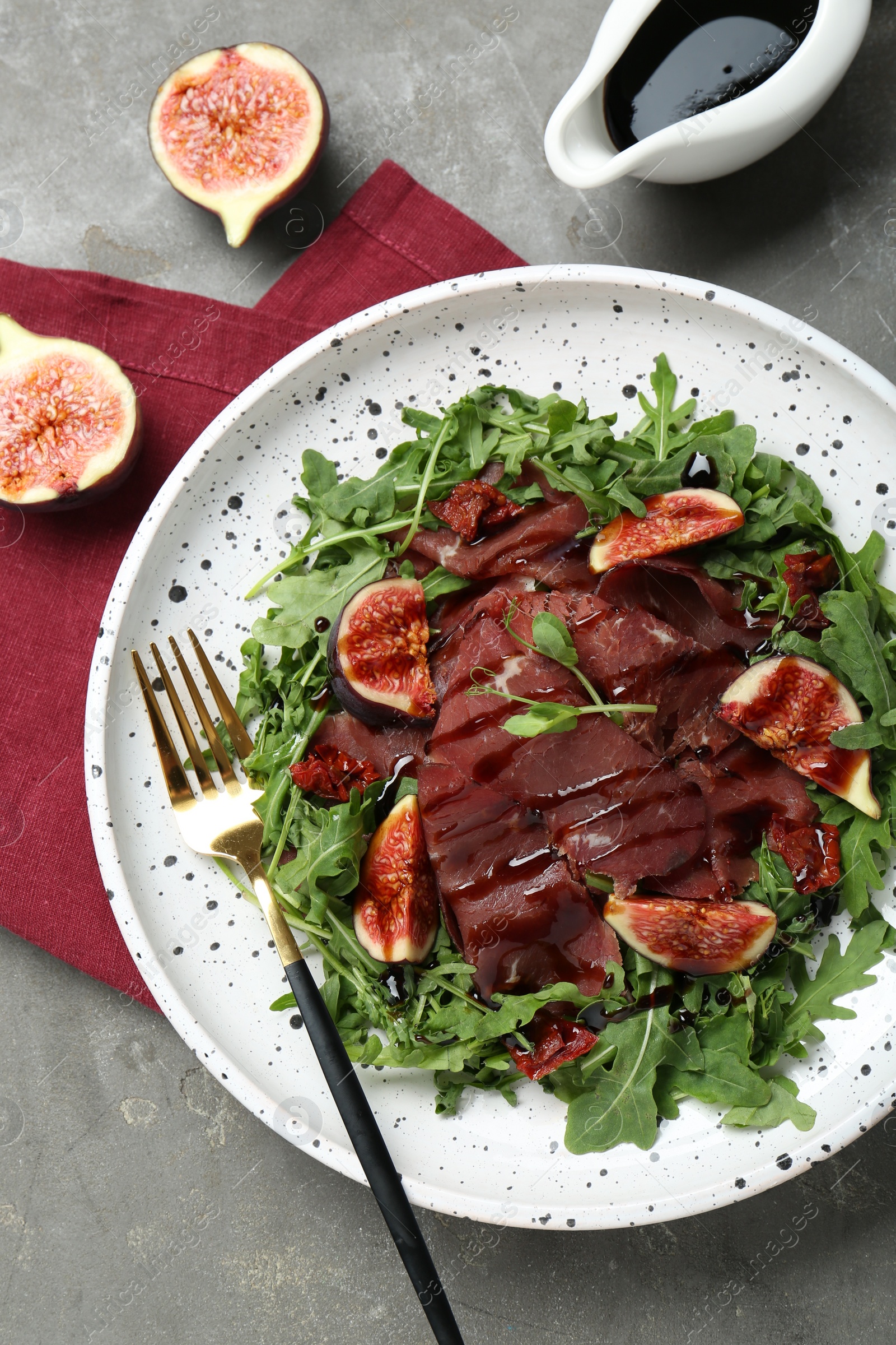 Photo of Plate of tasty bresaola salad with figs, sun-dried tomatoes, balsamic vinegar and fork on grey table, flat lay