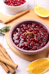 Photo of Tasty cranberry sauce in bowl and ingredients on white tiled table, closeup