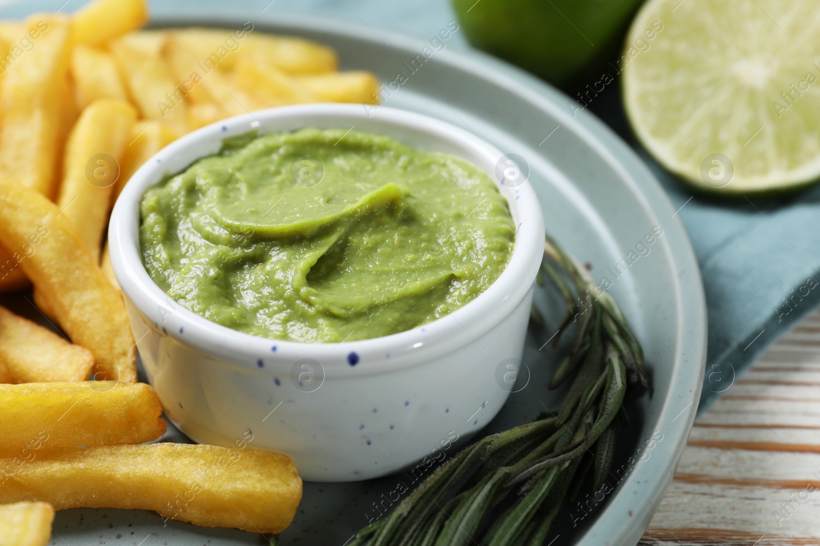 Photo of Plate with delicious french fries, avocado dip, lime and rosemary served on white wooden table, closeup
