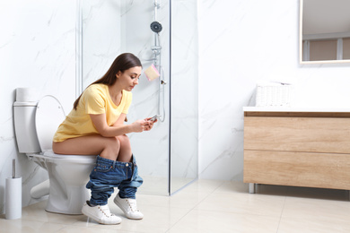 Woman with smartphone sitting on toilet bowl in bathroom