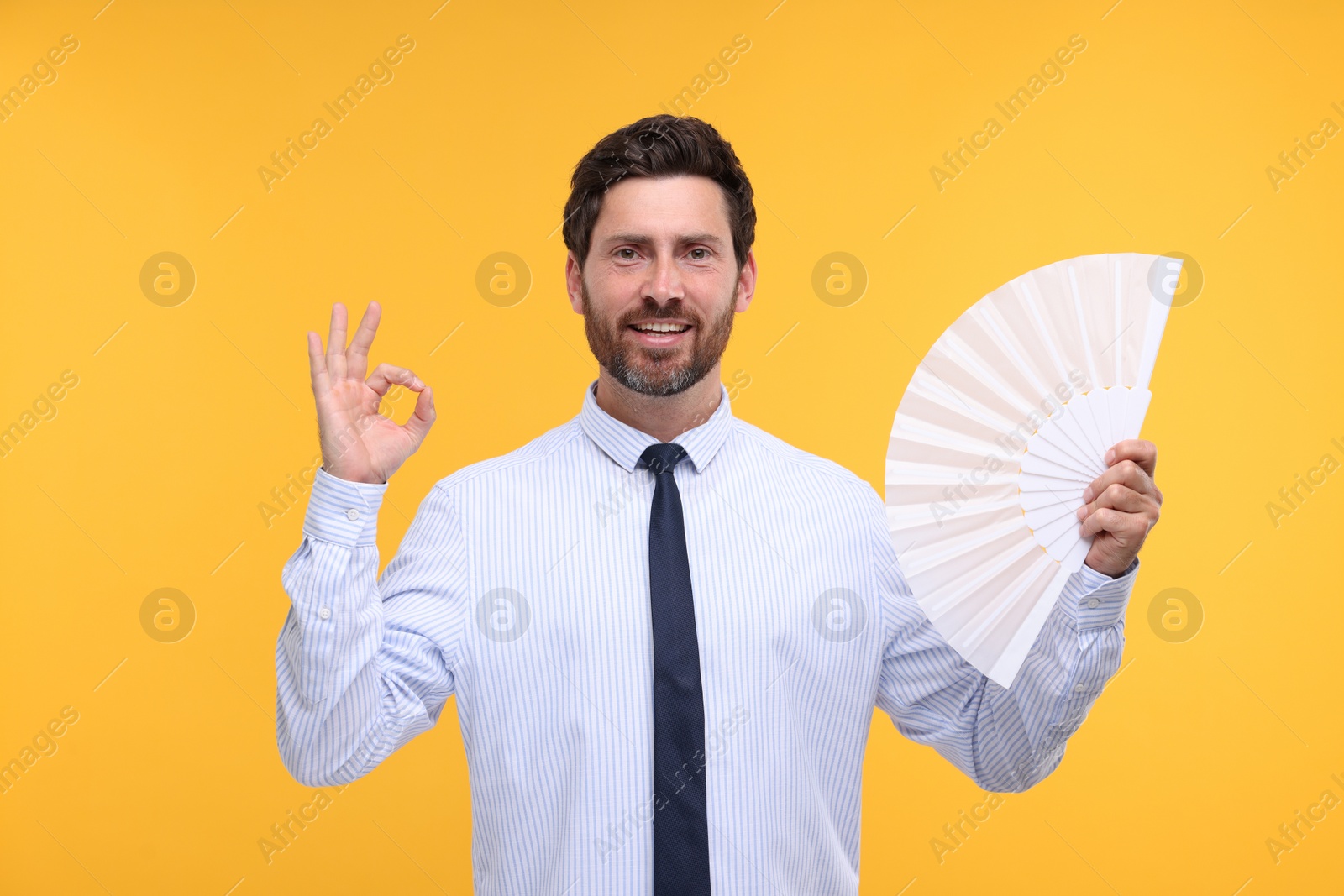 Photo of Happy man holding hand fan and showing ok gesture on orange background