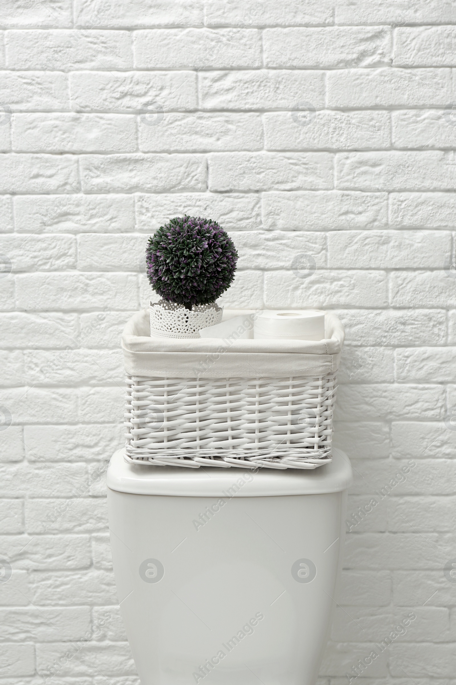 Photo of Basket with roll of paper and decorative plant on toilet tank near white brick wall. Bathroom interior