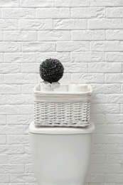 Basket with roll of paper and decorative plant on toilet tank near white brick wall. Bathroom interior