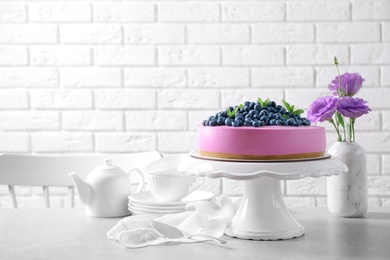 Photo of Stand with tasty blueberry cake and flowers on grey table against white brick wall. Space for text