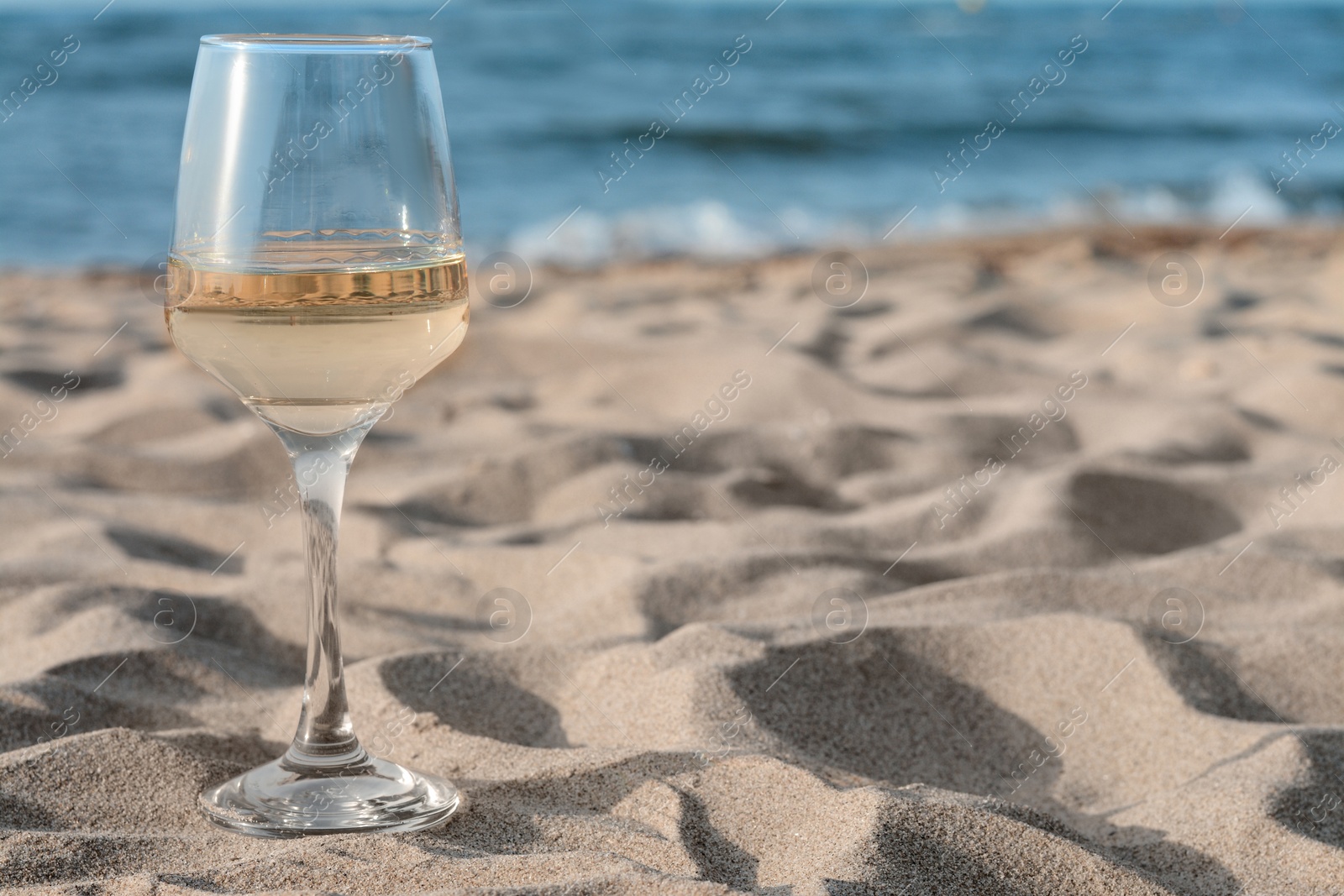 Photo of Glass of tasty wine on sand near sea, space for text