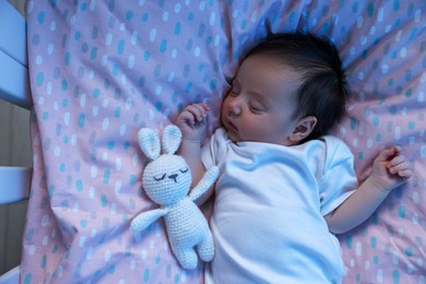 Cute newborn baby sleeping with toy bunny in crib at night, top view