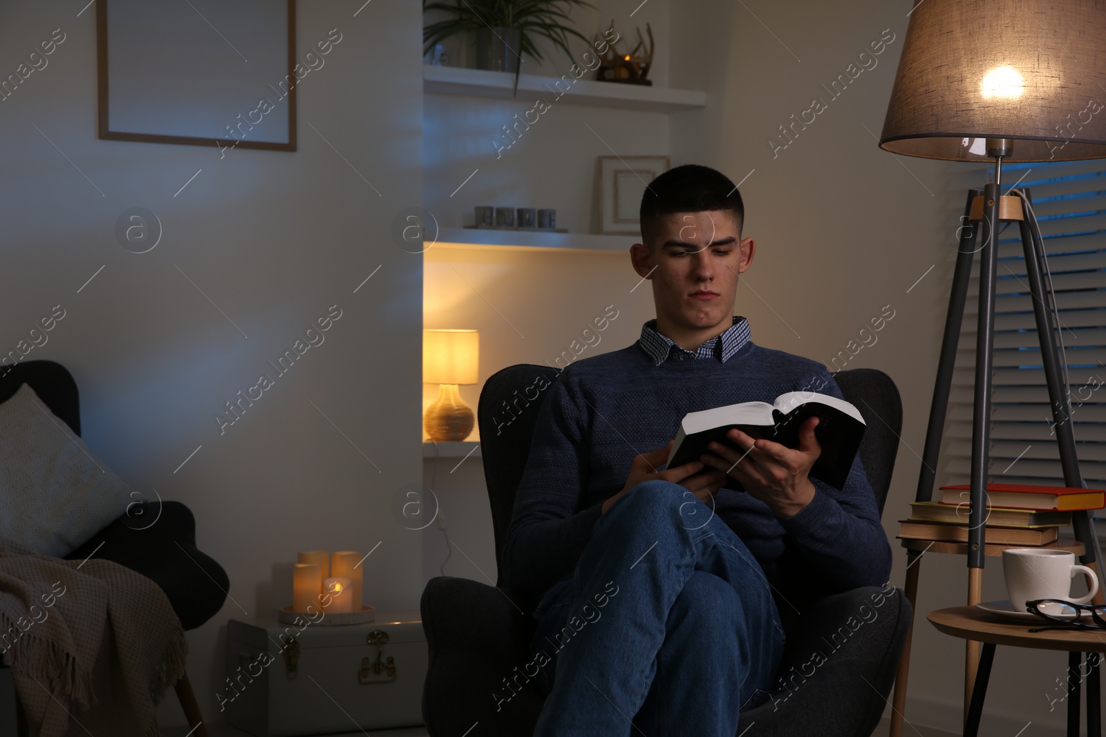 Photo of Young man with cup of drink reading book in cozy room at night