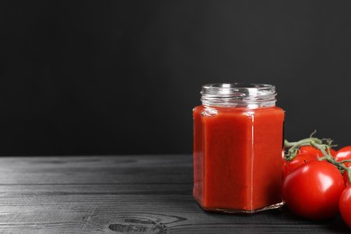 Photo of Delicious ketchup in jar and tomatoes on black wooden table, closeup with space for text. Tomato sauce